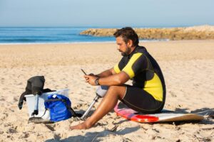 A man with a prosthetic leg uses a mobile phone while sitting on a surfboard at the beach.